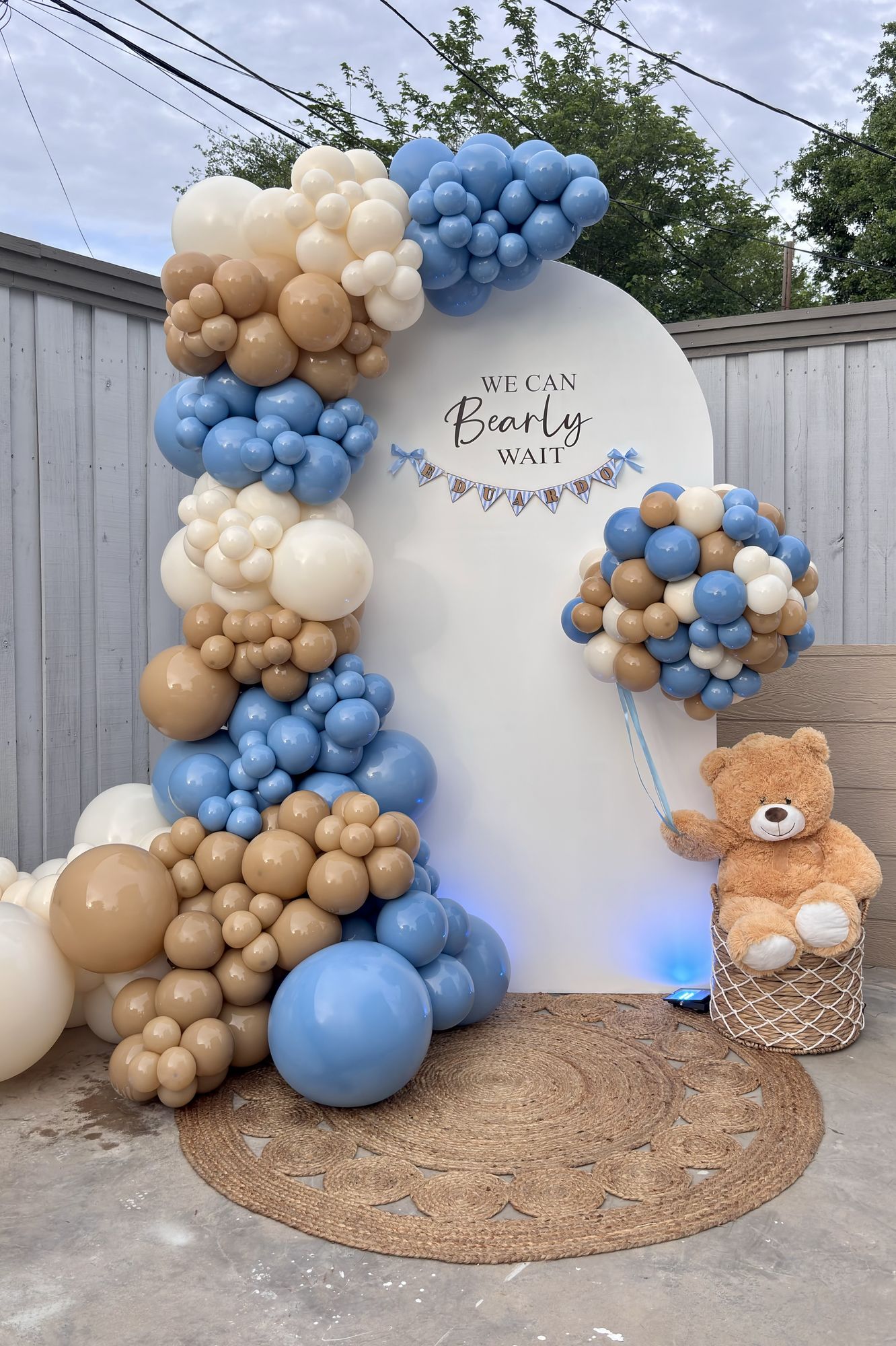 Blue, tan and cream balloon garland on a white arch backdrop with a teddy bear for a baby shower