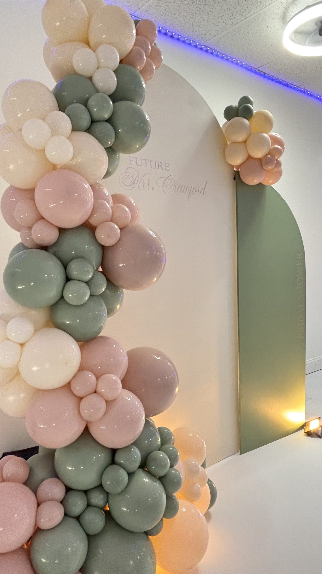 Sage green, blush and cream organic balloon arch beside a wall with gold bridal shower script